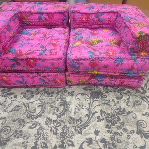 May include: Two matching pink upholstered armchairs with floral patterns in blue, red, and yellow. The chairs are placed on a gray patterned rug. The chairs have a low profile and a boxy shape.