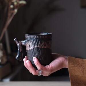 May include: A black and brown ceramic mug with a textured surface and a unique handle. The mug is being held in a hand.