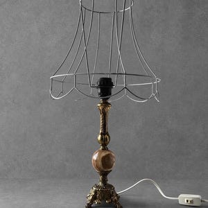 Può includere: Una lampada da tavolo con base decorativa in ottone, un piede in metallo ornato e smerlato e una sfera centrale in pietra marrone lucida. La lampada include un portalampada nero e un paralume in filo metallico aperto con bordo smerlato.
