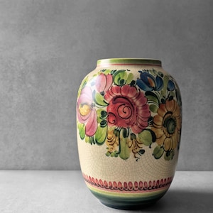 Może przedstawiać: Ceramiczny wazon o kremowej podstawie z wykończeniem w formie spękanego szkliwa. Zewnętrzna część jest ręcznie malowana w żywe motywy kwiatowe w odcieniach różu, żółci, błękitu i zieleni, z ozdobnym, falistym obramowaniem przy dolnej krawędzi.