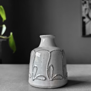 Może przedstawiać: Biały ceramiczny wazon z błyszczącym wykończeniem, szeroką, zaokrągloną podstawą i zwężającą się szyjką. Zewnętrzna powierzchnia ozdobiona jest powtarzającym się wzorem ciemnych, teksturowanych, ręcznie rysowanych motywów płatków lub liści.