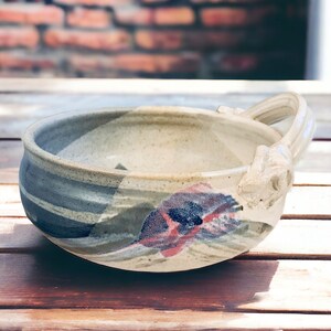 May include: A white ceramic bowl with a unique, sculpted handle. The bowl has a hand-painted design with shades of blue, gray, green, and pink.