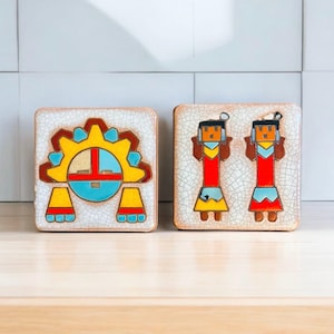 Puede incluir: Dos azulejos de cerámica con diseños de nativos americanos. El primer azulejo presenta un sol estilizado con una cara en amarillo, rojo, azul y blanco. El segundo azulejo presenta dos figuras en rojo, amarillo, azul y blanco con cabello negro y plumas. Ambos azulejos tienen un fondo blanco con un esmalte craquelado.