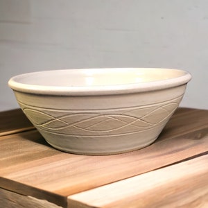 May include: A white ceramic bowl with a braided design around the rim. The bowl is sitting on a wooden surface.