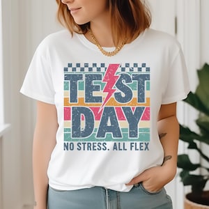 以下が含まれることがあります： 雷とカラフルなストライプで「テストの日」と書かれたレトロなグラフィックがプリントされた白いTシャツ。グラフィックの下のテキストには「ストレスなし。すべてフレックス。」と書いてあります。