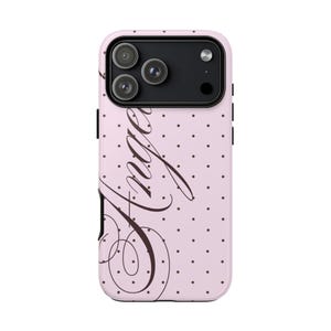 May include: A pink phone case with a black camera module and a polka dot pattern. The word "Angel" is written in a cursive font across the case.