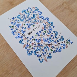 May include: A white card with a floral design in shades of blue, green, and red. The word "INVICTUS" is written in the center of the card. The card is on a wooden surface.