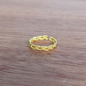 Puede incluir: Un anillo dorado con un diseño trenzado. El anillo está abierto en la parte superior, lo que permite ajustar la talla. El anillo está hecho de un material metálico brillante y se encuentra sobre una superficie de madera.