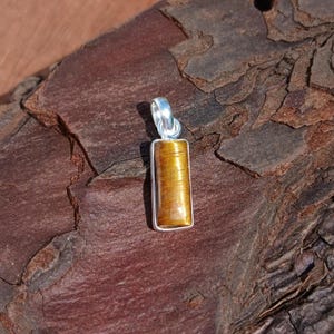Puede incluir: Un colgante rectangular con una piedra de ojo de tigre pulida engastada en un marco de metal plateado. El colgante tiene un lazo en la parte superior para una cadena. La piedra ojo de tigre muestra tonos marrón dorado y amarillo.