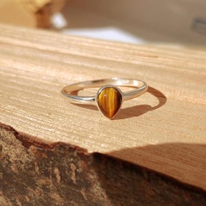 May include: A silver ring featuring a teardrop-shaped tiger's eye gemstone. The stone displays brown and gold stripes. The ring is displayed on a wooden surface, highlighting the jewellery's design and colour.