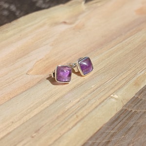 May include: A pair of silver stud earrings with square amethyst gemstones. The earrings are set against a wooden background.