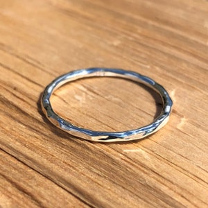 Puede incluir: Un anillo de plata con textura martillada. El anillo es circular y se encuentra sobre una superficie de madera. La superficie del anillo refleja la luz, creando reflejos y sombras. Es una joya sencilla y elegante.