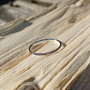 May include: A thin silver ring with a smooth, polished finish. The ring is lying on a wooden surface.