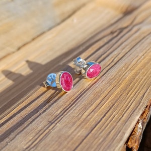 Könnte beinhalten: Ein Paar Ohrstecker aus Sterlingsilber mit ovalen, facettierten, leuchtend roten Edelsteinen. Die Ohrringe sind in einer silbernen Fassung gefasst und werden auf einer Holzoberfläche präsentiert, die die lebendige Farbe zeigt.