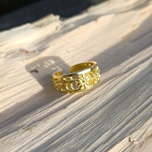 May include: A gold-coloured adjustable toe ring with a floral design. The ring features an open design with cut-out flowers and leaves. The ring is displayed on a light-coloured wooden surface.