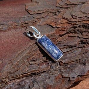 May include: A rectangular blue gemstone pendant with a silver-colored metal frame and bail. The gemstone has a textured appearance with streaks of lighter blue. The pendant is displayed on a textured, brown wooden surface.