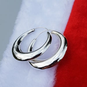 Peut inclure: Une paire de boucles d'oreilles créoles en argent avec une surface lisse et arrondie. Les boucles d'oreilles sont grandes et ont un style contemporain.