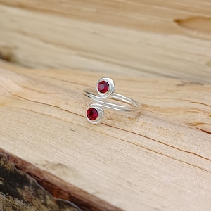 Może przedstawiać: Srebrny pierścionek z dwoma czerwonymi kamieniami szlachetnymi osadzonymi w obrączce. Pierścionek jest zaprojektowany z zakręconą obrączką.