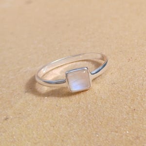 Op de afbeelding: Een zilveren ring met een vierkante, lichtgeleurde edelsteen. De ringband is dun en gepolijst, en de edelsteen is gezet in een vierkante zetting. De ring wordt getoond op een lichtbruin oppervlak.