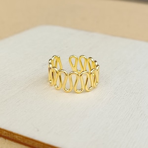 Pode incluir: Anel ajustável de ouro com um design ondulado e orgânico. O anel é feito de uma única peça de fio que é torcido e moldado em uma série de laços.