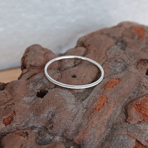 May include: A simple silver band ring, shown on a textured brown surface.