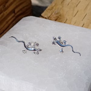 Può includere: Un paio di orecchini a forma di lucertola in argento con una coda curva. Gli orecchini sono su una superficie bianca.