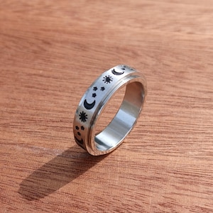 May include: A silver-colored ring with a celestial design featuring moons, stars, and dots. The ring is set against a wooden surface, showcasing its metallic sheen and intricate details. The ring's design is etched in black.