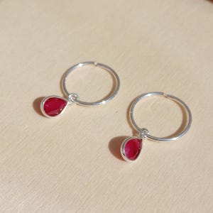 Op de afbeelding: Een paar zilveren oorringen, elk met een druppelvormige, dieprode edelsteenhanger. De oorbellen worden gepresenteerd op een lichtbruin oppervlak, met schaduwen.