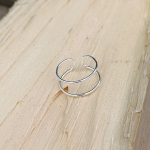 May include: A silver double band ring with a simple design. The ring is made of two thin bands of metal that are joined together at the top. The ring is shown on a wooden surface.