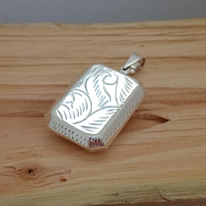 Könnte beinhalten: Silberner Anhänger mit einem gravierten Blumenmuster. Das Medaillon ist rechteckig und hat einen Klappdeckel.