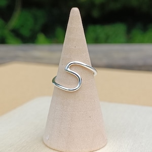 Può includere: Un anello d'argento a forma di onda, esposto su un supporto per anelli in legno.