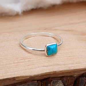 Op de afbeelding: Een zilveren ring met een vierkante, turquoise steen. De smalle band is gepolijst en de turquoise steen heeft een heldere blauwgroene kleur. De ring ligt op een houten oppervlak.