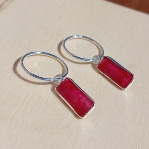 Op de afbeelding: Een paar zilveren oorringen met rechthoekige, dieprode edelsteenhangers. De edelstenen zijn gezet in een zilveren rand. De oorbellen worden gepresenteerd tegen een neutrale achtergrond.