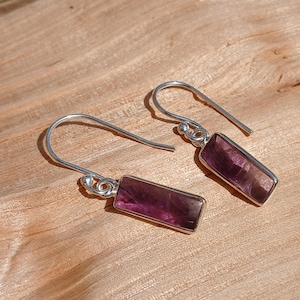 May include: Silver drop earrings featuring rectangular, purple gemstones. The earrings are displayed on a light brown wooden surface. The gemstones have a translucent quality, and the earrings are designed with a hook closure.