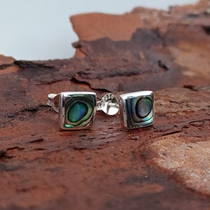 May include: Square sterling silver stud earrings with iridescent abalone shell inlays. The abalone shell features a swirling pattern of blues, greens, and purples. The earrings are set against a natural wood background.