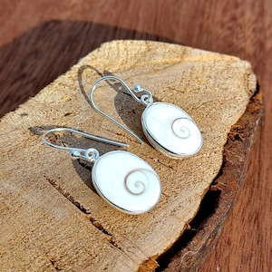 Peut inclure: Une paire de boucles d'oreilles en argent avec un motif de coquille blanche. La coquille a un motif en spirale au centre.