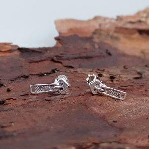Peut inclure: Une paire de boucles d'oreilles en argent en forme de tirette de fermeture éclair. Les boucles d'oreilles sont petites et délicates, et elles présentent un design réaliste de tirette de fermeture éclair.