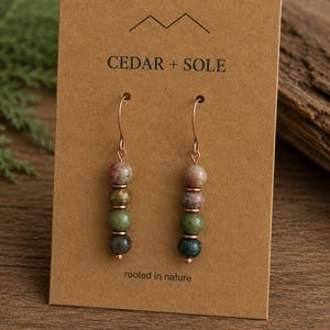 Könnte beinhalten: Ein Paar Ohrhänger auf einer braunen Karte mit der Aufschrift CEDAR + SOLE und rooted in nature. Jeder Ohrring besteht aus vier gestapelten, kugelförmigen Perlen in Rosa-, Beige- und Grüntönen, die an einem kupferfarbenen Metallhaken hängen.