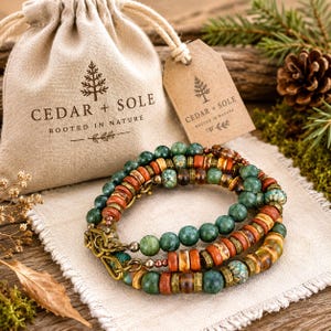 May include: A multi-strand bracelet featuring green, brown, and orange beads with metallic spacers, positioned in front of a beige fabric drawstring pouch and a brown paper tag, both printed with the text CEDAR + SOLE ROOTED IN NATURE.