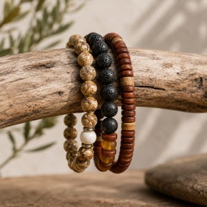 May include: Three beaded bracelets arranged together. One features speckled tan and brown stone beads with a single white bead. The second consists of black porous lava rock beads. The third is made of small, cylindrical dark brown wooden beads.