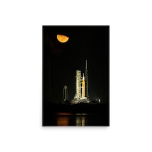 May include: A nighttime photograph of a rocket on a launchpad, illuminated by bright lights. The rocket is white and orange, with a water tower and other structures nearby. A large, orange half-moon is visible in the dark sky above the launch site.