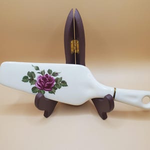 May include: White ceramic cake server with a purple rose design. The server is on a wooden stand.