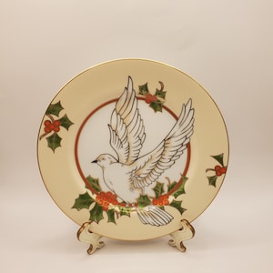 May include: A white porcelain plate with a gold rim features a hand-painted design of a white dove flying with outstretched wings. The dove is surrounded by green holly leaves with red berries.