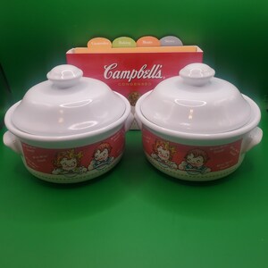 May include: Two white ceramic soup bowls with lids, featuring a red and white design with a cartoon image of two children eating soup. The bowls have the text "M'm! M'm! Good!" on the side. The bowls are in front of a red box with the text "Campbell's Condensed" on it.