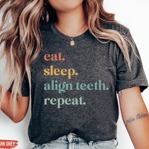 May include: Heather dark grey t-shirt with the text "eat. sleep. align teeth. repeat." in colorful letters.