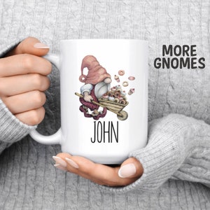 May include: White ceramic coffee mug with a pink gnome pushing a wheelbarrow full of donuts and cupcakes. The mug has the name "JOHN" printed on the bottom.