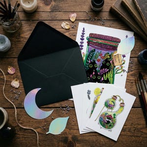 May include: A collection of whimsical art prints and stationery on a wooden surface. The set includes illustrated cards with cats, plants, and potion bottles, a black envelope, and iridescent moon and leaf shapes. The artwork has a fantasy theme.