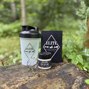May include: A clear plastic shaker bottle with a black lid, a small jar, and a black card, all bearing the "ELITE" logo. The shaker has a metal whisk inside. The items are arranged on a tree stump against a blurred green background.