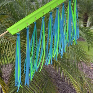May include: A neon green decorative item with blue and green ribbons hanging from it. The ribbons are attached to a green bar with metal rings. The item is set against a backdrop of palm trees and a cloudy sky.