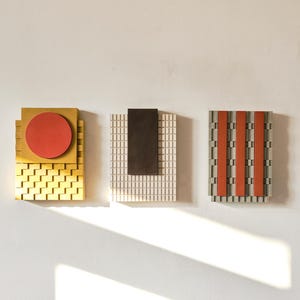 May include: Three abstract wall art pieces with geometric designs. The first features a red circle on a gold square, the second a dark brown rectangle on a grid, and the third has red stripes. All are mounted on a white wall.